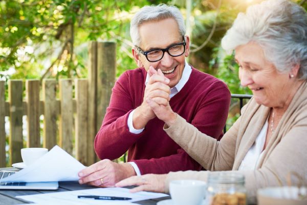 Un couple de personnes âgées en train de sourire tout en consultant des documents, assis à une table dans un jardin, avec des tasses de café à côté d’eux.