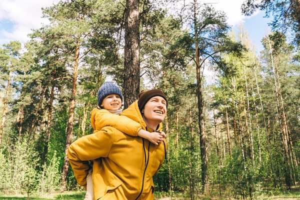 Un homme, chaudement vêtu, se promène en forêt par une belle journée, en tenant son fils par les 2 bras dans son dos.