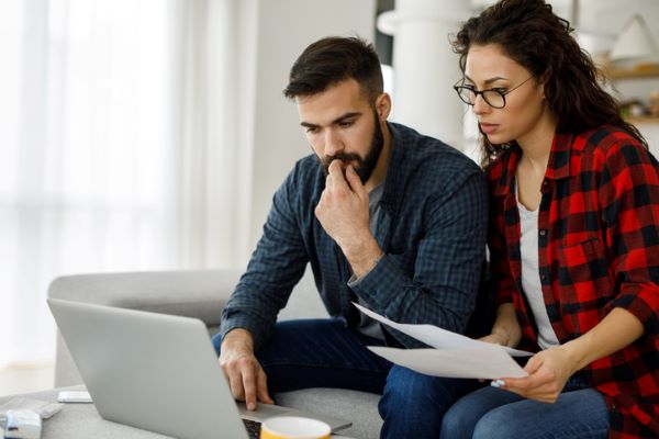 Un jeune couple, assis côte à côte sur un canapé, passe en revue leur compte sur un ordinateur portable posé devant eux.